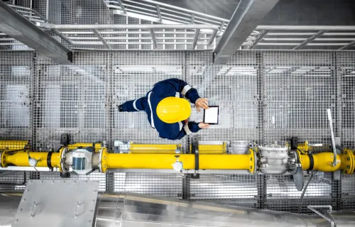 Refinery worker with yellow hardhat walking by gas pipes